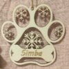 Décoration de Noël en bois clair en forme de patte de chien, gravée du prénom de votre animal (comme "Simba" sur le modèle). Le design hivernal est sublimé par de délicats flocons de neige découpés au laser et une zone en forme d'os pour le texte. C'est le souvenir parfait ou le cadeau idéal pour tout amoureux des chiens.